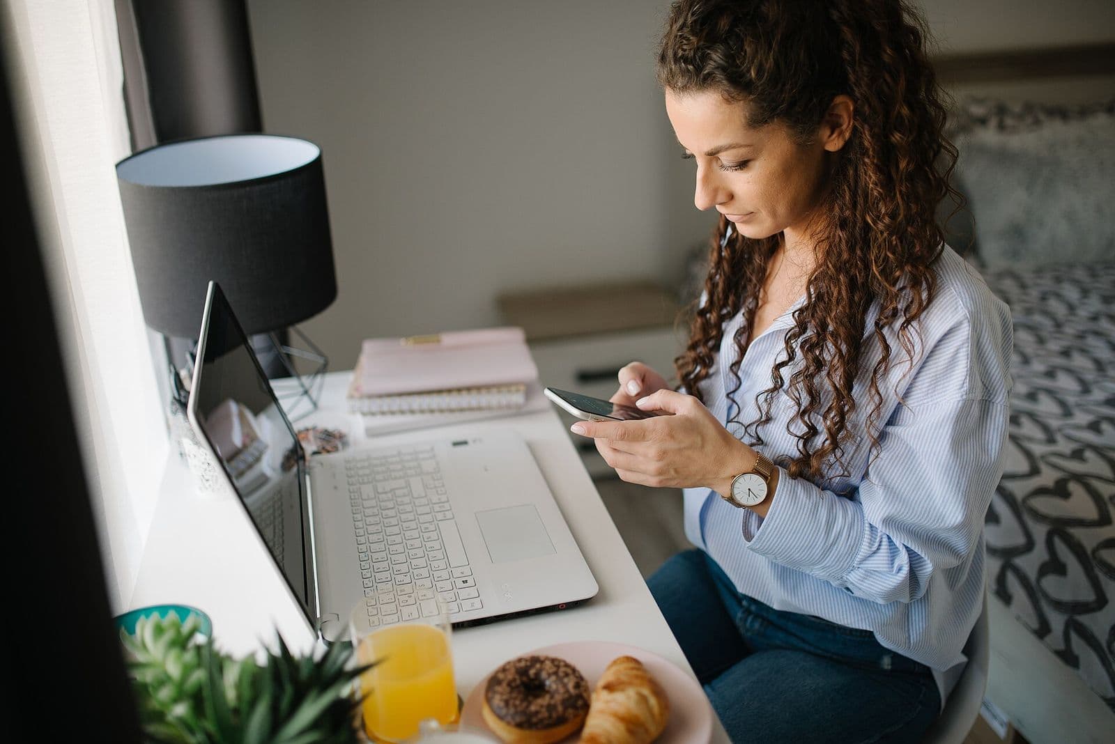 Woman browsing on her smartphone at a desk with a laptop and breakfast