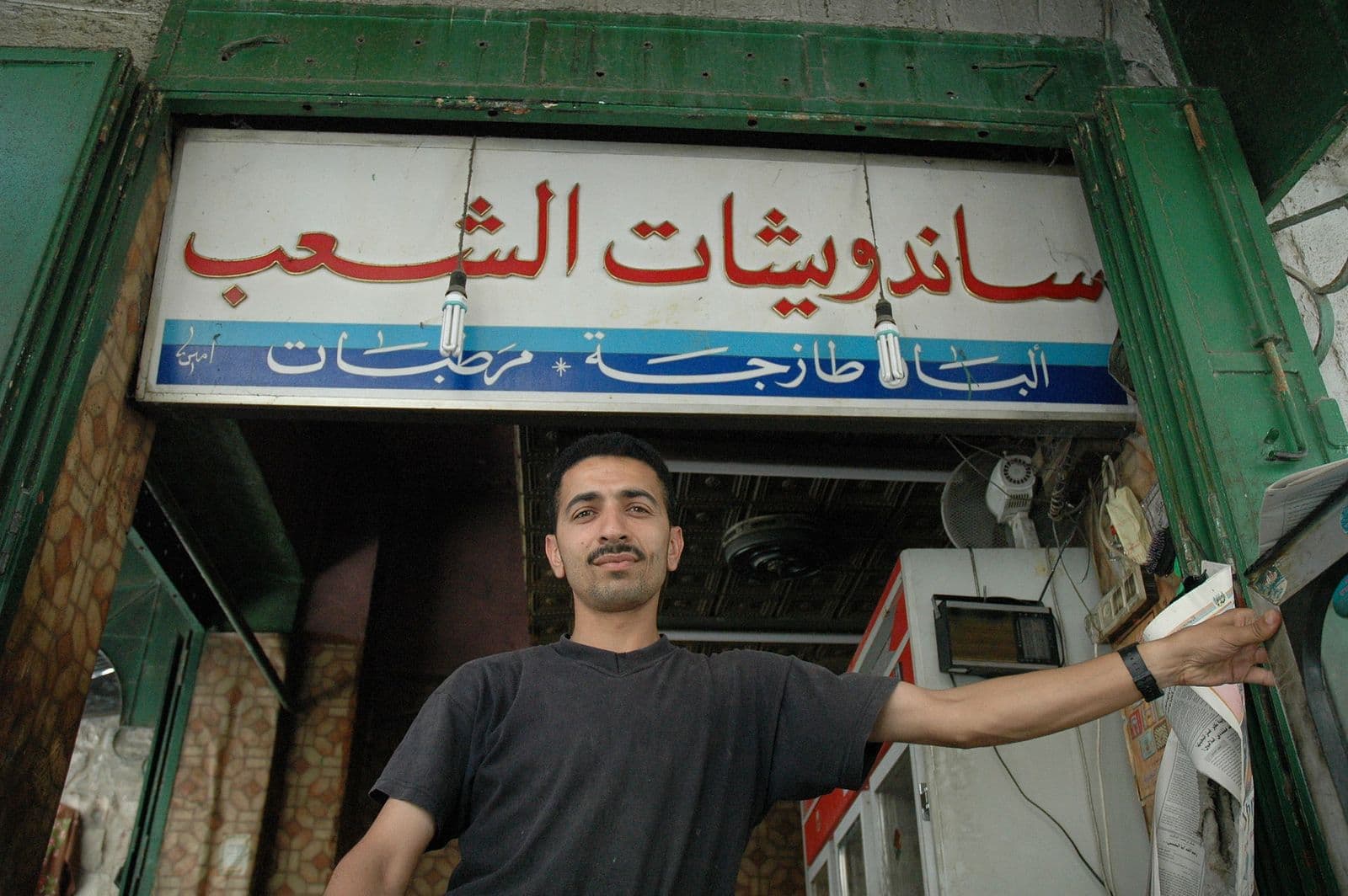 Owner standing in the doorway of his small sandwich shop under an Arabic sign